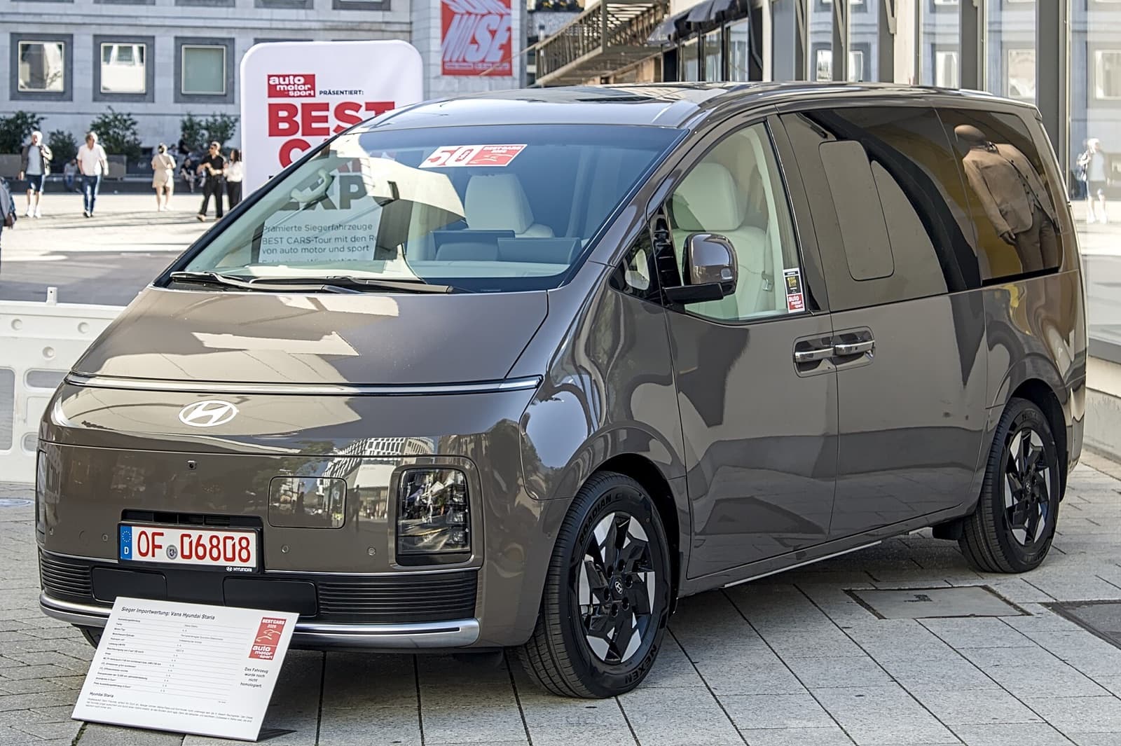 Bronze Hyundai STARIA Electric van on a Stuttgart street, front three-quarter view with closed grille and German dealer plate