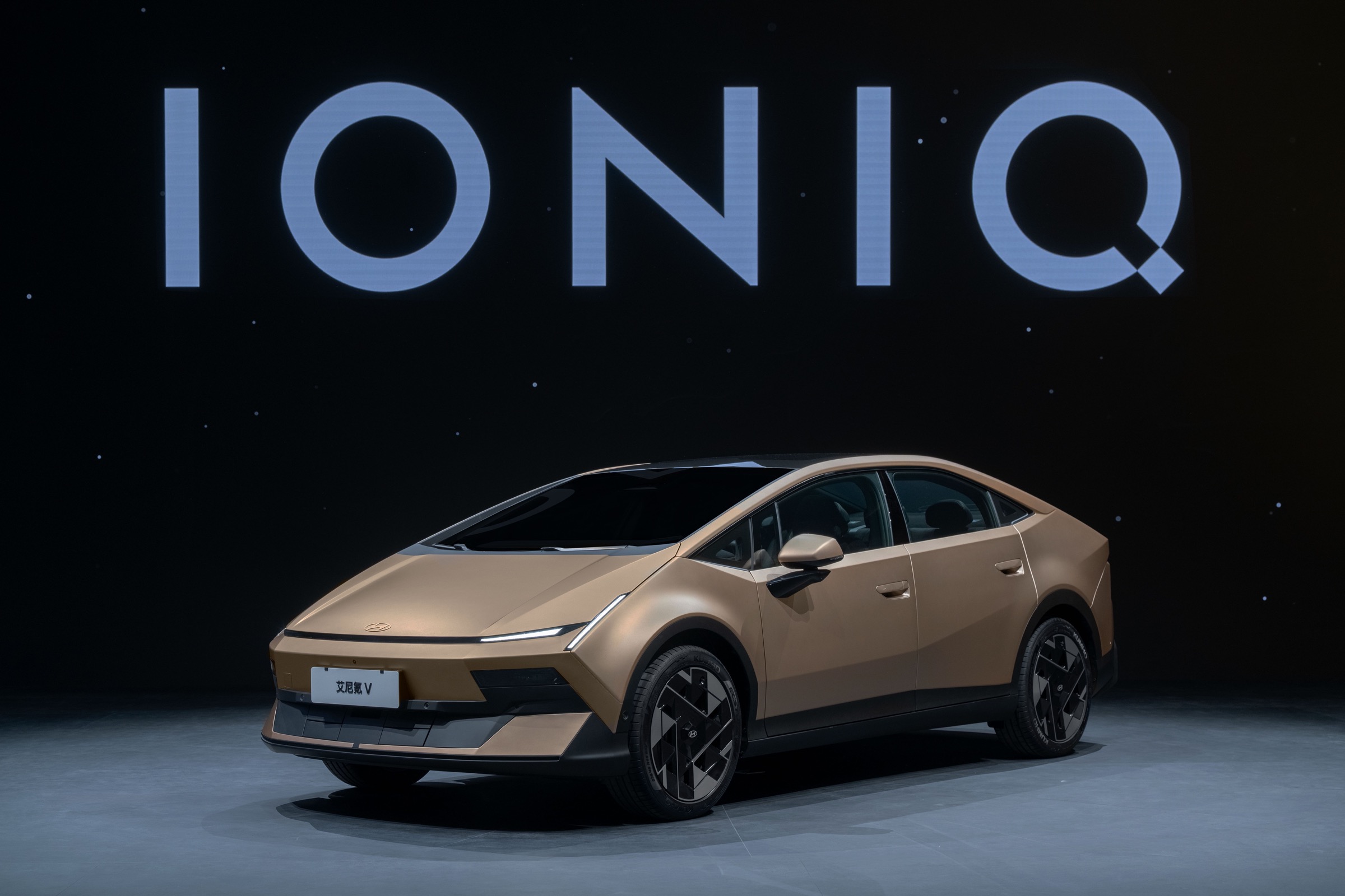 Gold Hyundai Ioniq V fastback sedan on a studio floor under an IONIQ backdrop