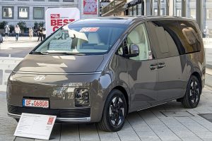 Bronze Hyundai STARIA Electric van on a Stuttgart street, front three-quarter view with closed grille and German dealer plate
