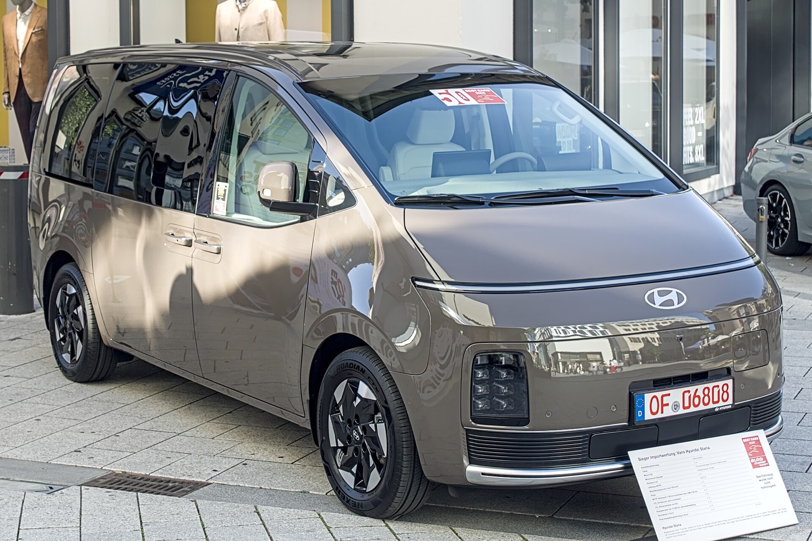 Bronze Hyundai STARIA Electric van, front three-quarter view on a Stuttgart street, closed grille and German dealer plate